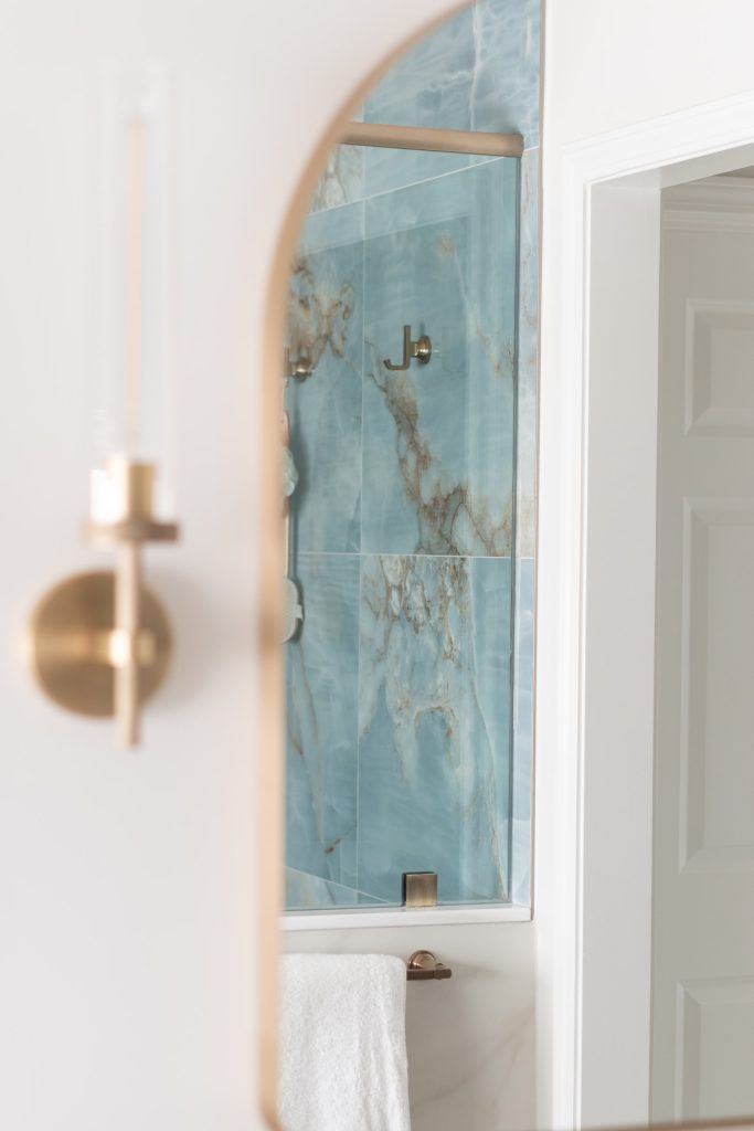 Brushed-brass sconce in the foreground with an arched mirror reflecting the veined blue onyx shower behind it.