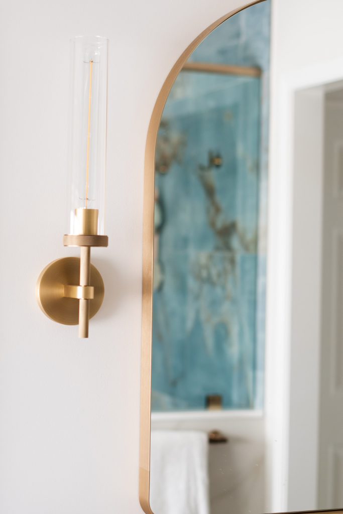 Brass-and-glass cylinder sconce in the foreground with an arched mirror reflecting the veined blue onyx shower.