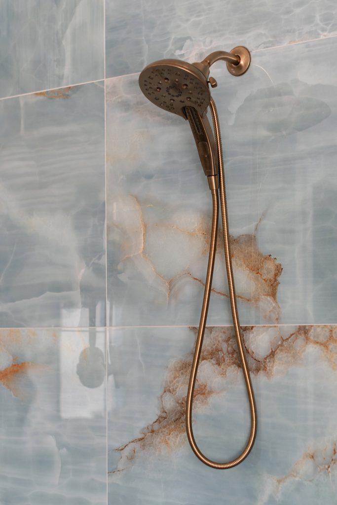 Brushed-bronze handheld shower head on a coiled hose against blue onyx tile with vivid amber veining.