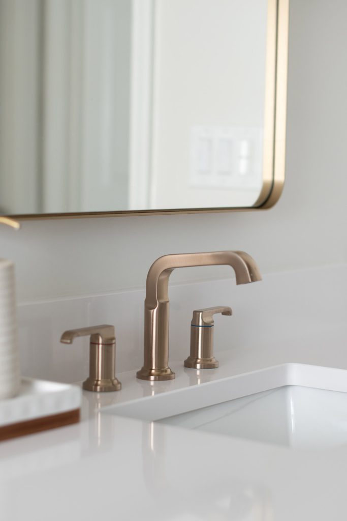 Close-up of a champagne-bronze widespread faucet on a white quartz counter beneath a brass-framed mirror.