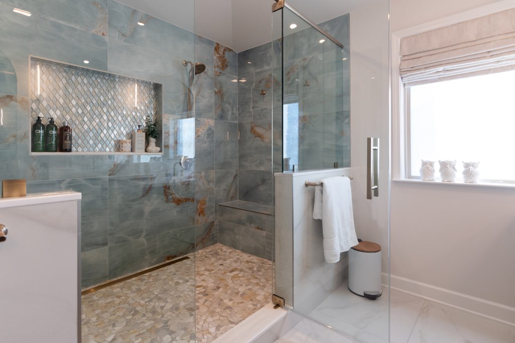 Onyx stone shower with a lit diamond mosaic niche, a bench, and a pebble floor, beside a window with a linen roman shade.