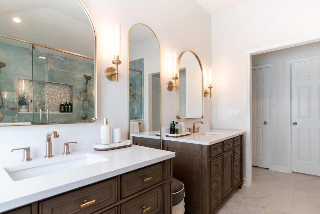 Master bath with two dark-wood vanities, white quartz counters, arched brass mirrors, and the blue onyx shower reflected behind.