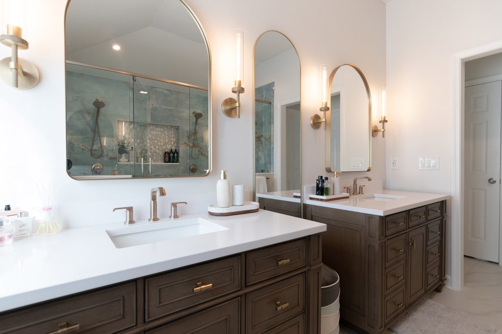 Twin dark-wood vanities with white quartz tops and arched brass mirrors that reflect the blue onyx shower and its diamond mosaic niche.