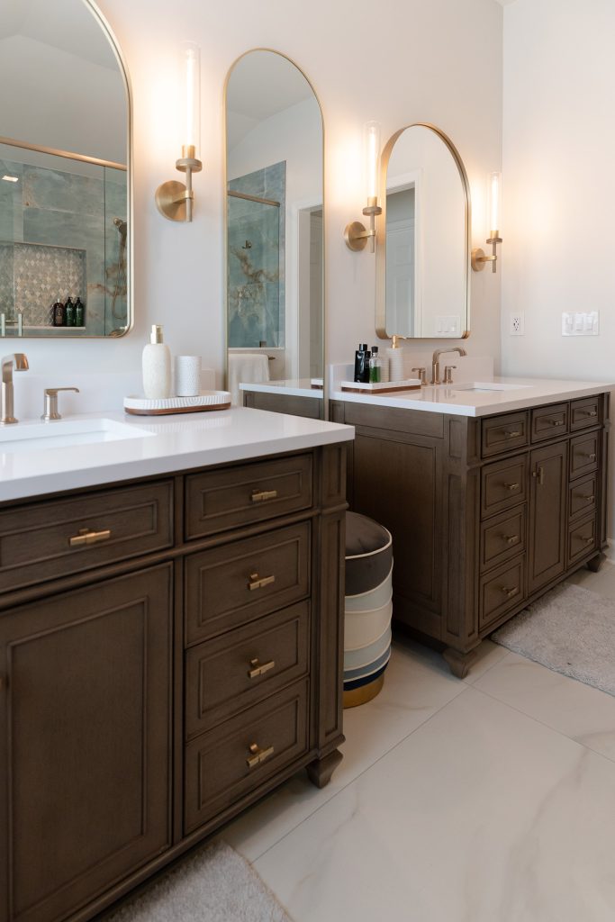 Two dark-wood vanities in a row with white quartz tops, arched brass mirrors, and a striped ottoman on marble-look flooring.