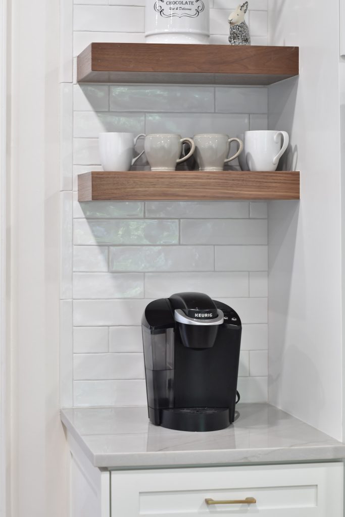 Coffee station with walnut floating shelves, gray mugs, a Keurig, and white subway tile on a marble-look counter in Alpharetta.