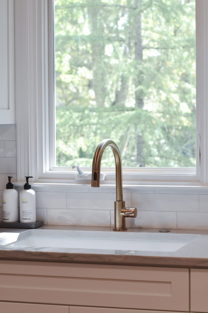 Brushed-gold gooseneck faucet over an undermount sink at a window, with white subway tile and a stone-edged counter in Alpharetta.