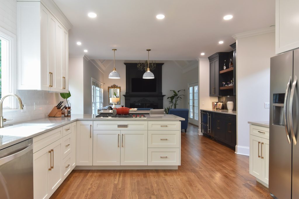 Bright white Alpharetta kitchen with a window over the sink, peninsula cooktop, gray pendants, and a dark living-room fireplace.