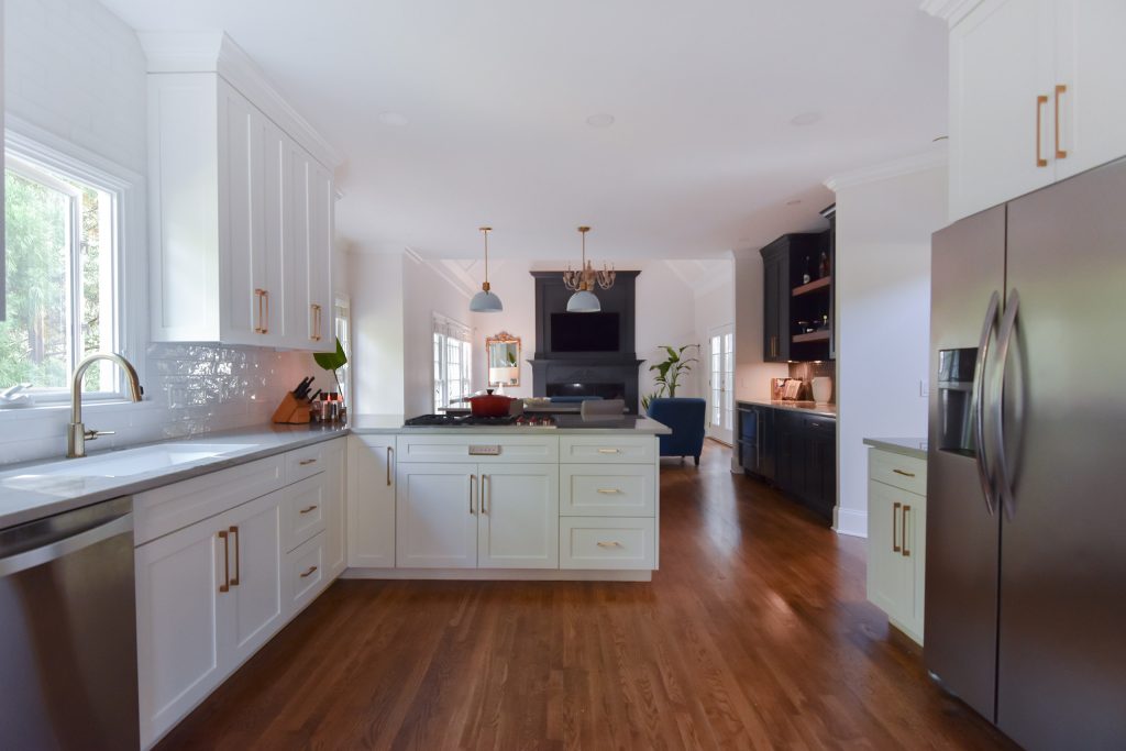 Wide white Alpharetta kitchen with oak floors opening to a living room with a dark fireplace and TV, and a navy bar at right.