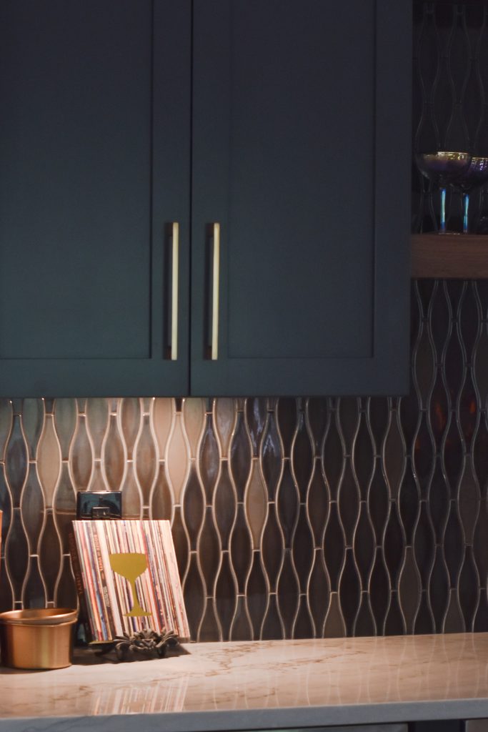 Navy bar cabinet with a gold pull and iridescent tile above a veined marble counter, with coupe glasses on a walnut shelf in Alpharetta.