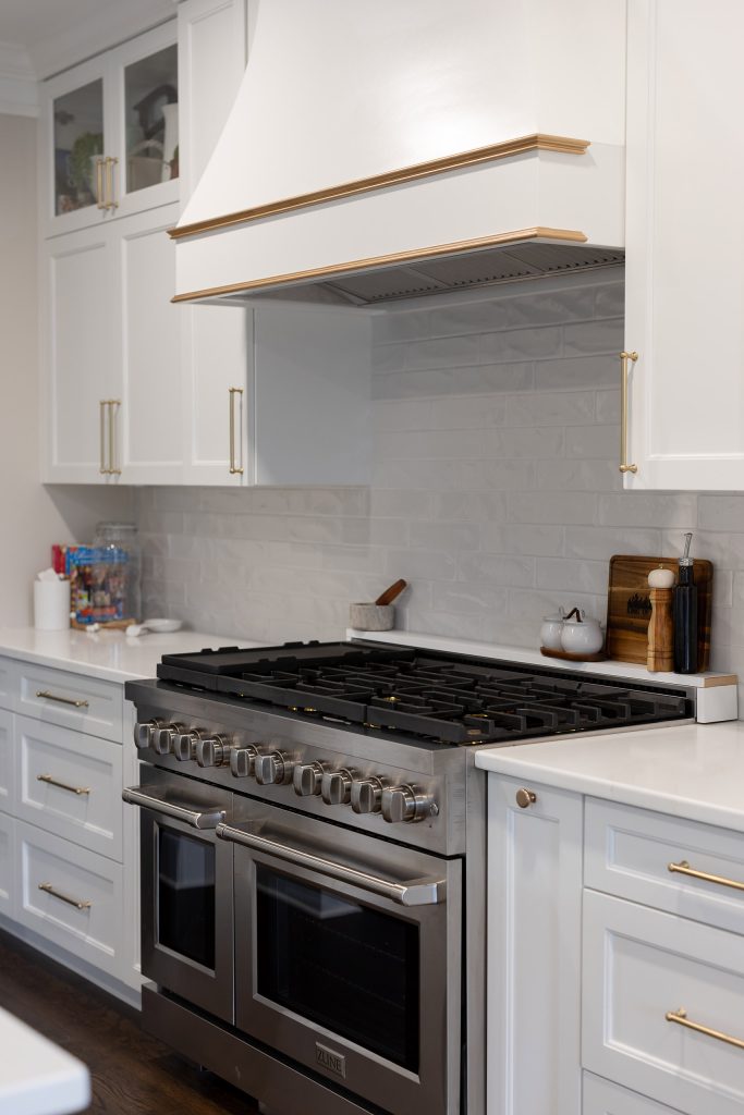 White plaster range hood with gold banding over a stainless pro range and glossy gray subway backsplash.