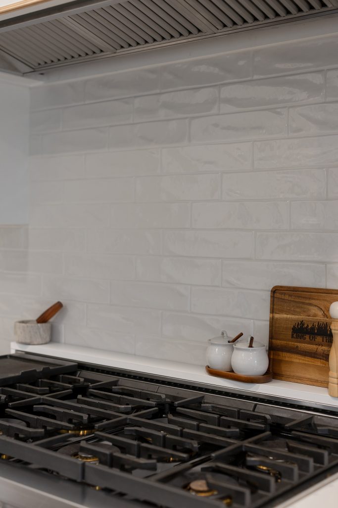 Stainless pro range with black cast-iron grates and gold burner accents against glossy gray tile and white cabinets.