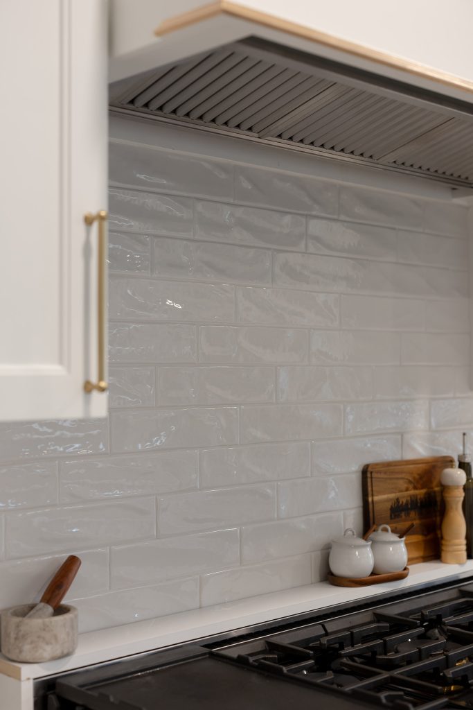 Glossy gray handmade-look backsplash tile beneath a stainless hood insert, with quartz shelf, mortar and spice jars.
