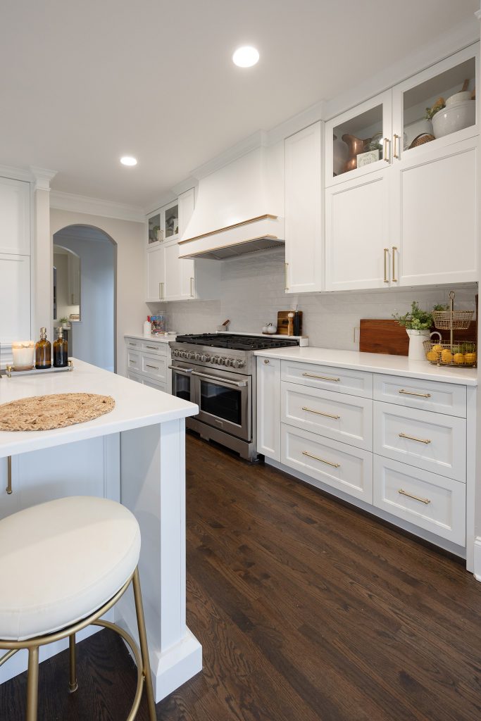 White cabinet run with brushed-gold hardware, stainless range, and a white leather barstool on a gold base at the counter.