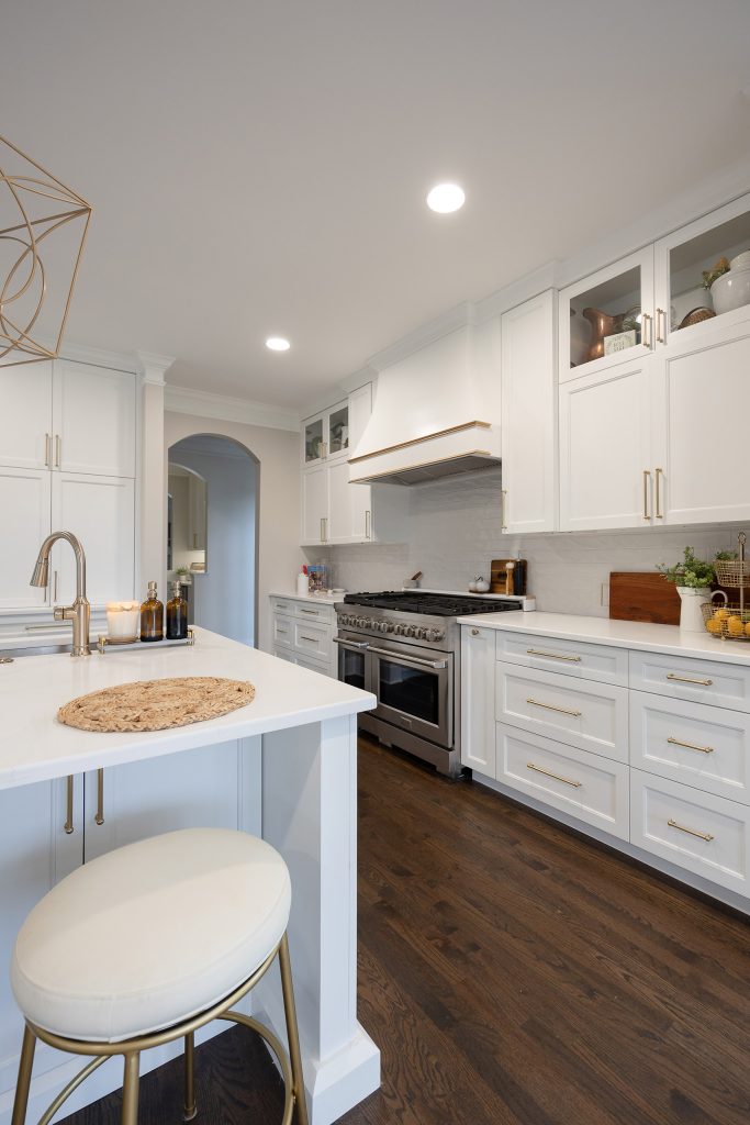 White kitchen with a gold-cushioned barstool at the island, stainless range, glass-front uppers, and dark oak floors.