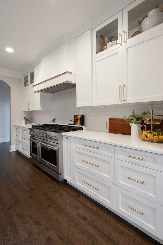 White shaker range wall with brushed-gold pulls, glossy gray backsplash tile, and a wire basket of lemons on quartz counters.