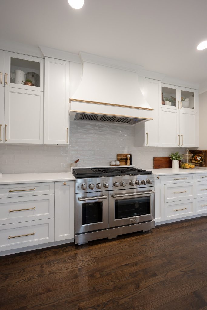 Stainless range under a white hood with gold-trimmed banding, flanked by white cabinets and a glossy gray tile backsplash.