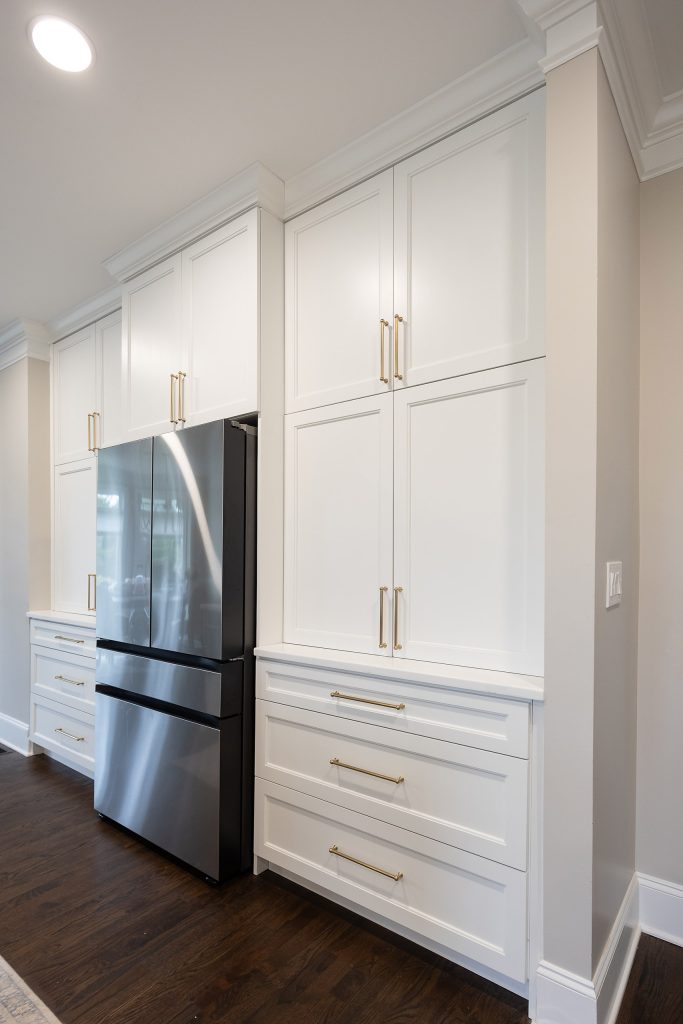 Stainless refrigerator built into floor-to-ceiling white shaker cabinets with brushed-gold bar pulls and dark hardwood floors.