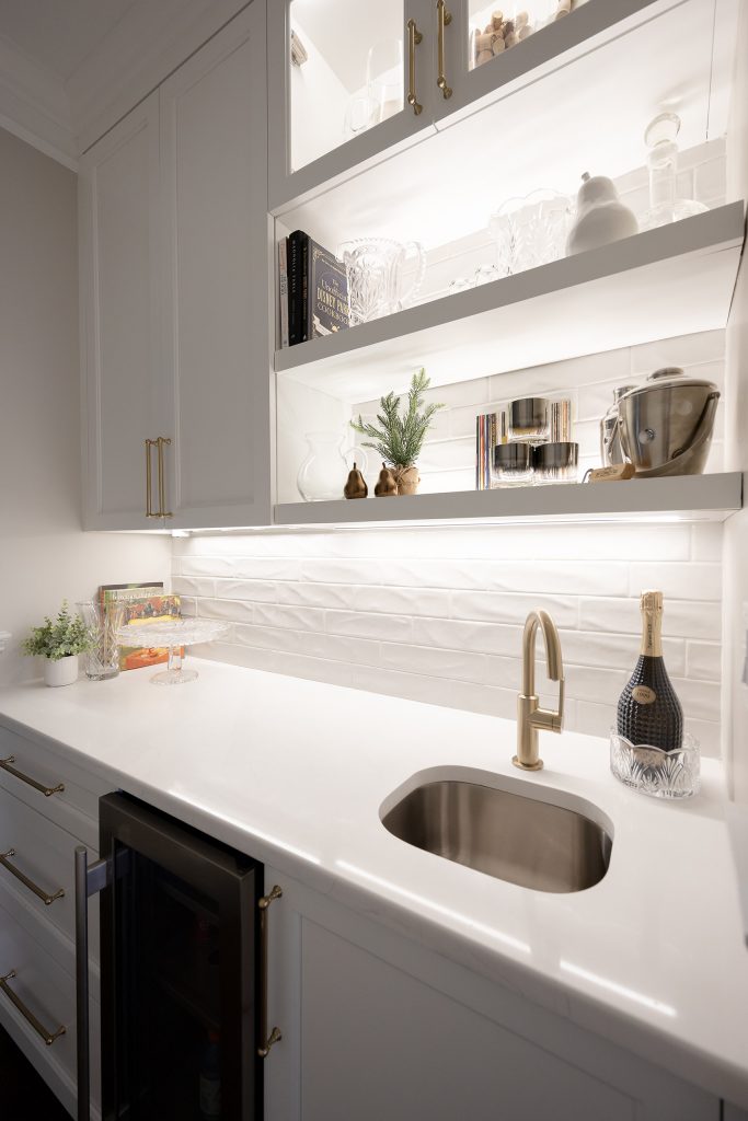 Butler's pantry open shelving with glassware and cookbooks, gold bar faucet, undermount sink, and glossy white tile.