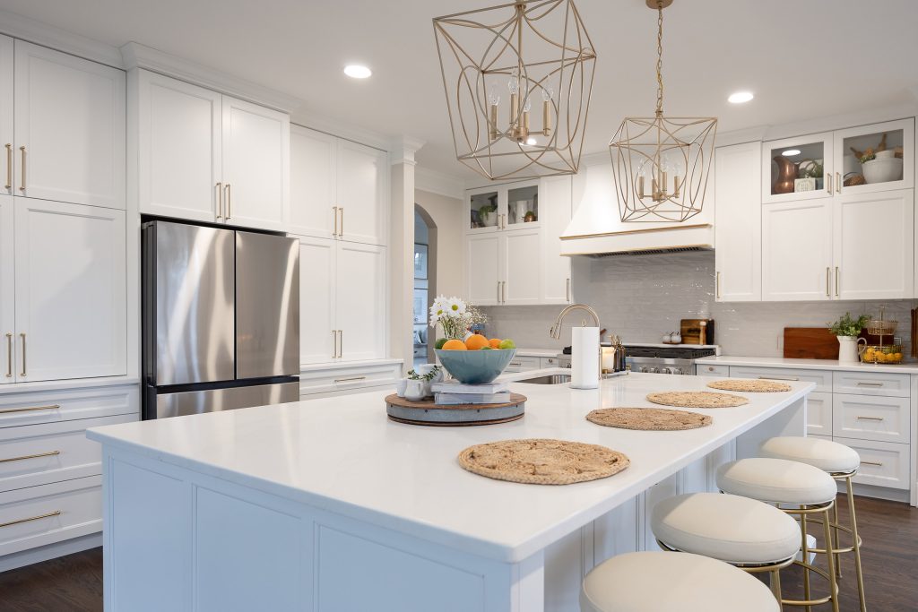 White quartz island with cream-and-gold barstools, stainless fridge, glass-front cabinets, and twin gold pendants.