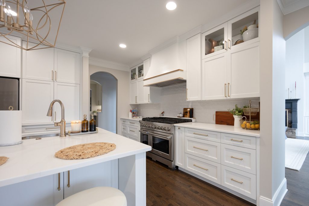 White kitchen run with stainless range and gold-trimmed hood, glass-front uppers, opening to a bright living room.