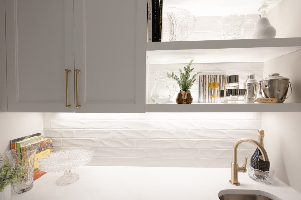 Butler's pantry with open lit shelves, glossy white tile, gold hardware, a bar sink, and a built-in wine fridge.