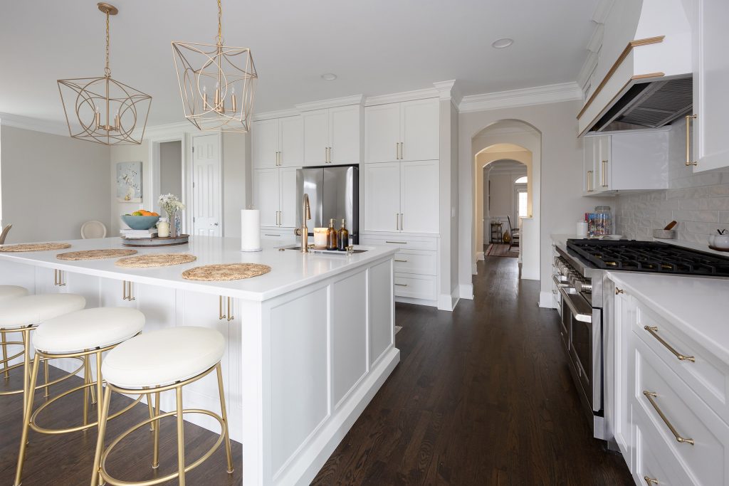 Open white-and-gold kitchen with a big island, cream-and-gold barstools, gold cage pendants, and French doors to a porch.