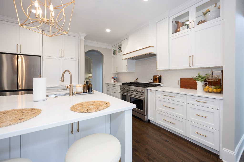End of a pale-blue island in white quartz with gold pulls, cream barstools, and a stainless range on the far wall.