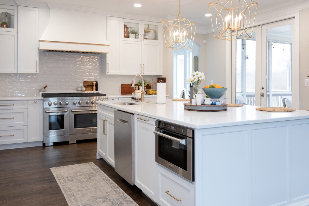 Wide white kitchen with a soft-blue island base, built-in microwave, gold pendants, and stainless appliances on dark oak.