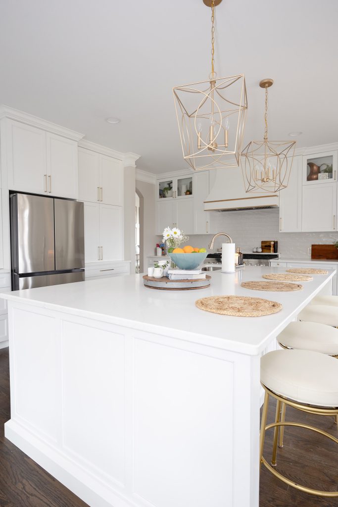 Tall white quartz island with round gold-based barstools, jute placemats, and gold cage pendants against white cabinetry.