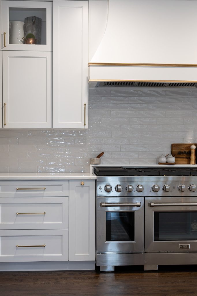 Stainless ZLINE range beneath a gold-trimmed white hood, set into white shaker cabinets with glossy gray backsplash.