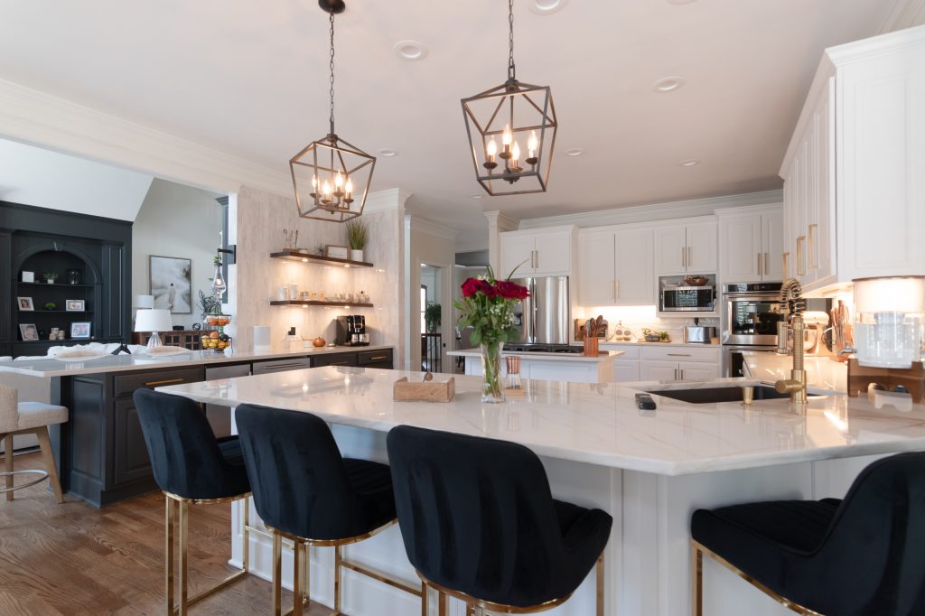 Open white kitchen with a large quartz island, black velvet and gold barstools, and lantern pendants over hardwood floors.