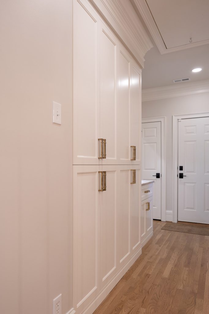Hallway with floor-to-ceiling white cabinets, aged-brass hardware, and white oak flooring toward a paneled door.