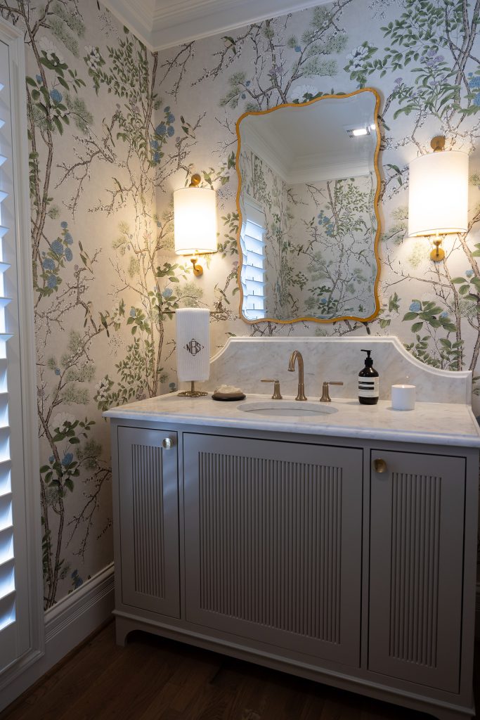 Powder room with a grey reeded vanity, marble counter, gold mirror, and a shuttered window on chinoiserie walls.