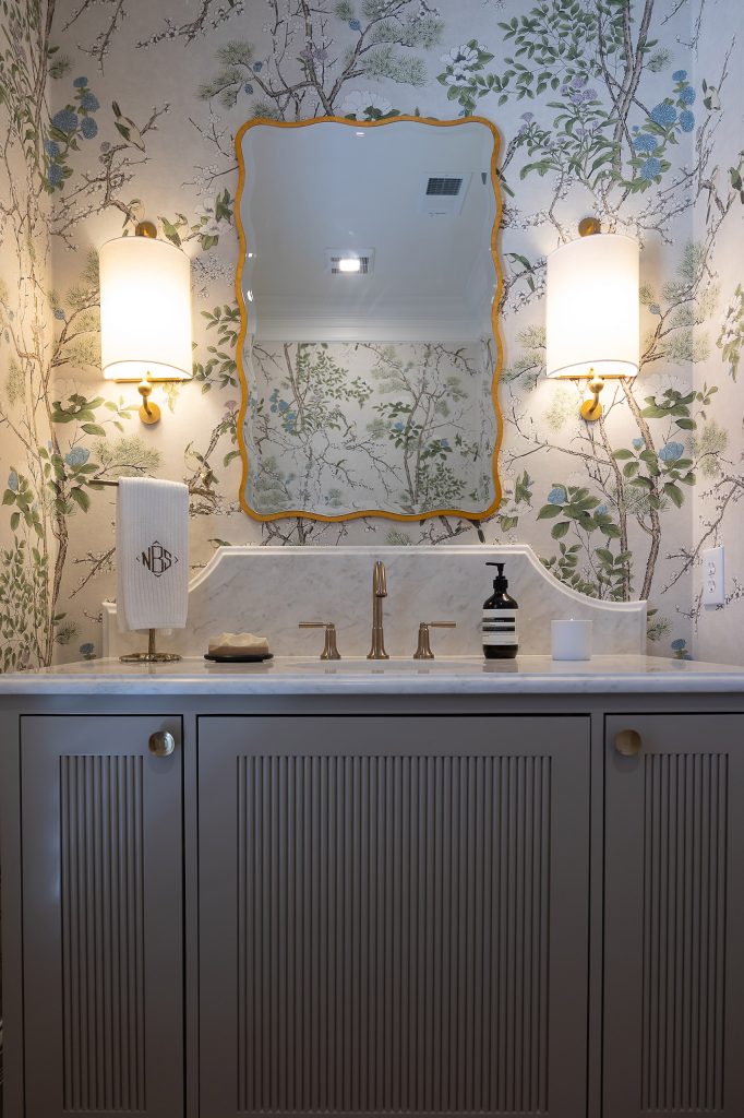 Grey reeded vanity with a marble top and brass faucet below a gold wavy mirror on chinoiserie wallpaper.