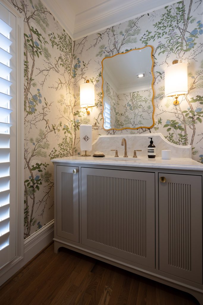 Powder room with a grey reeded vanity, marble top, gold wavy mirror, and lit brass sconces on chinoiserie walls.