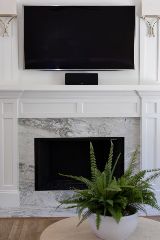 White paneled fireplace with a veined quartzite slab surround, a mounted flat-screen TV, and a fern.