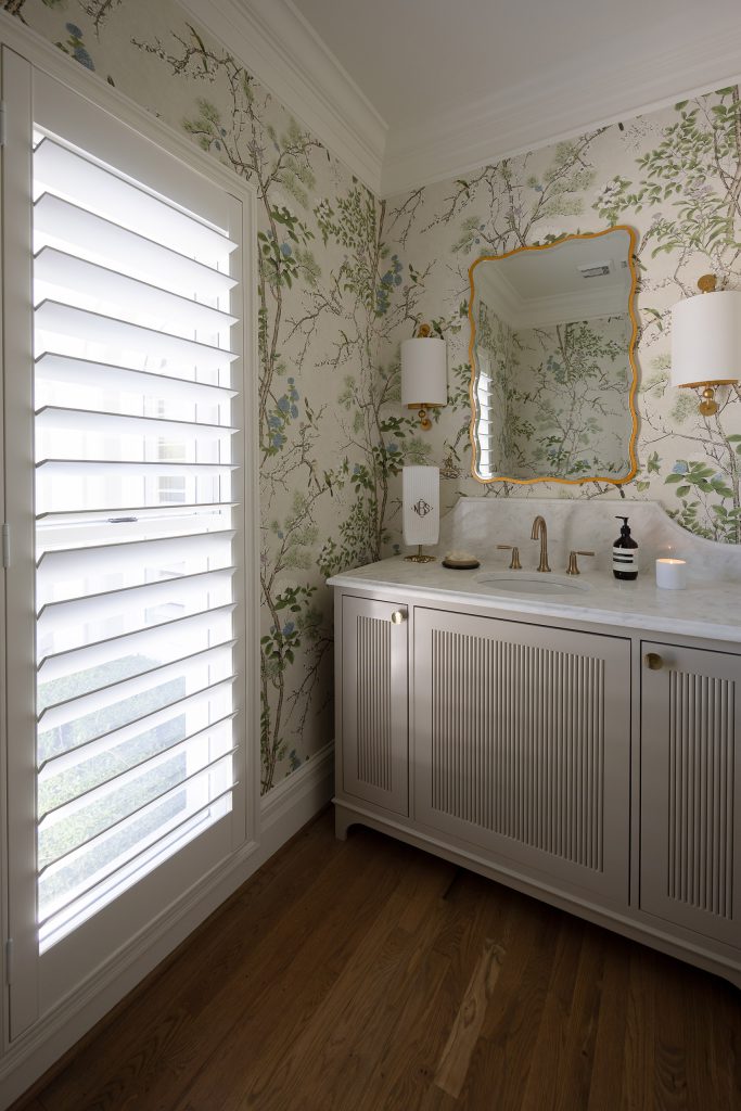 Powder room with chinoiserie wallpaper, a greige reeded vanity, marble top, and a plantation-shuttered window.