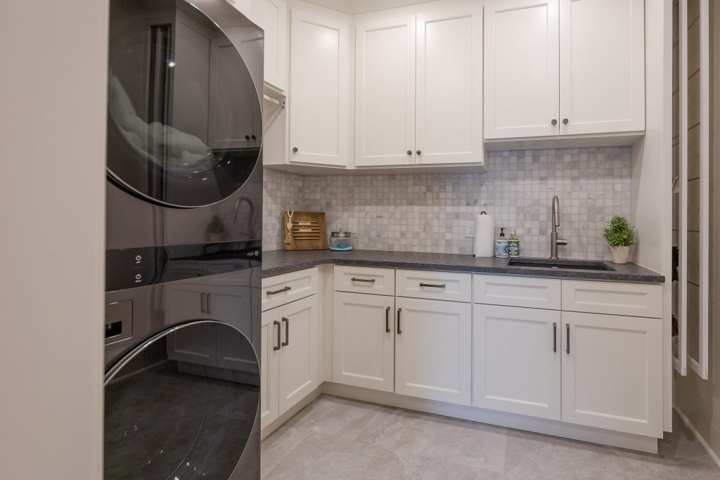 Laundry room with white shaker cabinetry, a marble mosaic tile backsplash, dark granite countertop, and a stacked washer and dryer.