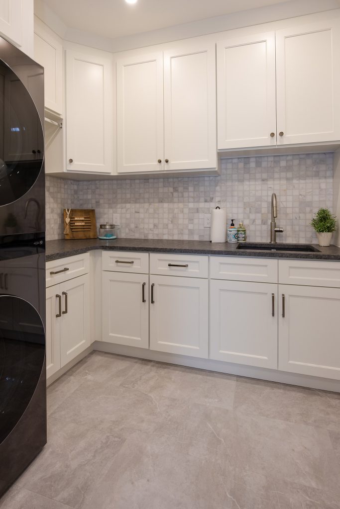 Laundry room with a black washer-dryer tower, white shaker cabinets, marble mosaic tile, and a stainless sink.