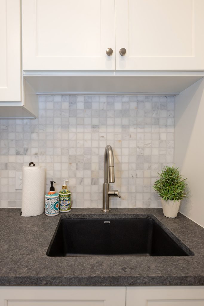 Laundry sink with white cabinets, a marble mosaic backsplash, a black composite sink, and dark granite counter.