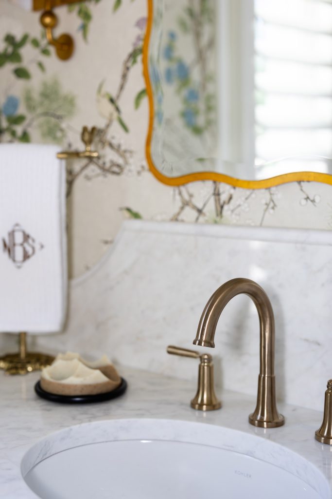 Aged-brass widespread faucet on a marble vanity with chinoiserie wallpaper and a monogrammed hand towel.