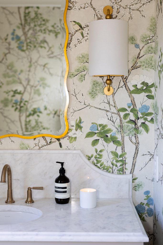 Powder room with green chinoiserie wallpaper, a gold wavy-edge mirror, marble counter, and brass faucet.