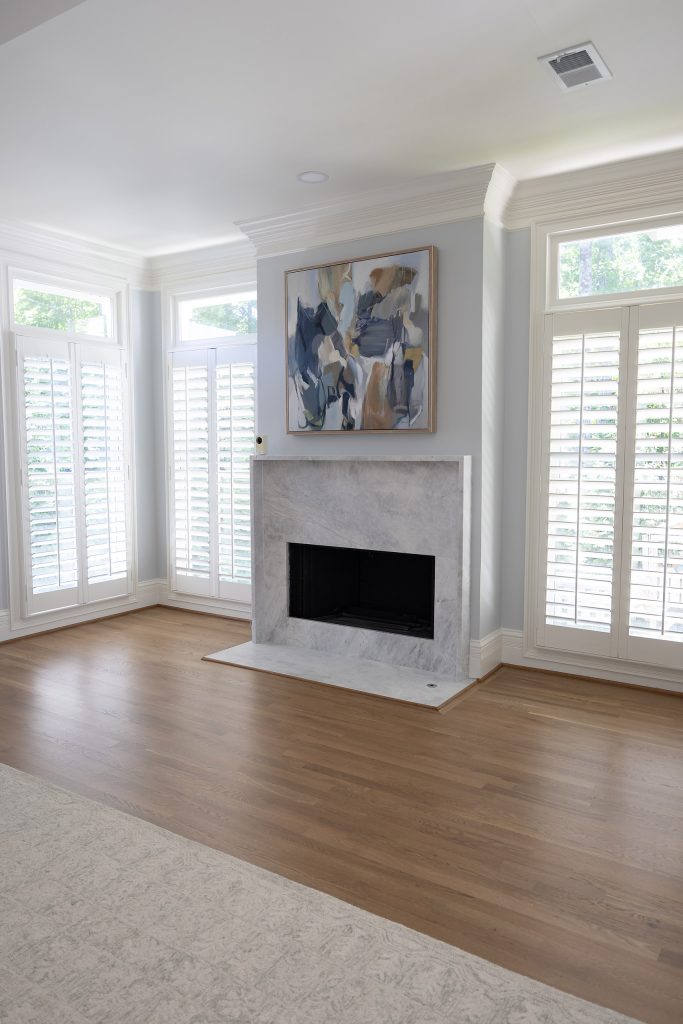 Primary bedroom with a veined stone-slab fireplace, abstract art, and shuttered windows over hardwood floors.