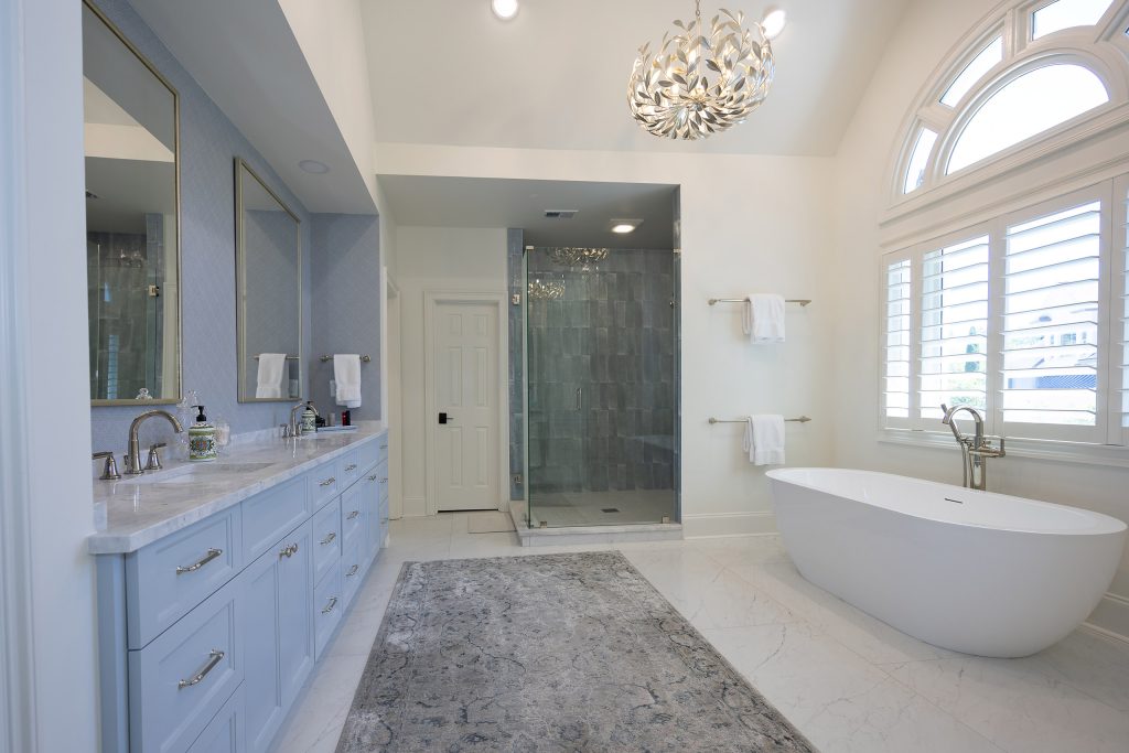Master bath with a grey-blue tiled glass shower, freestanding tub, arched window, and a white door.