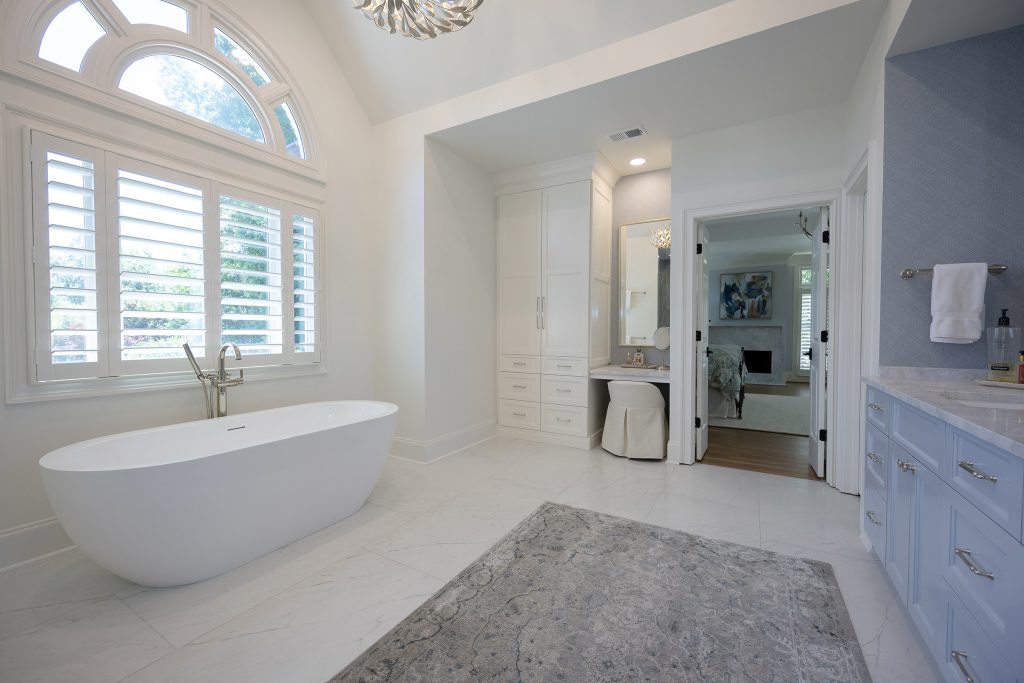 Wide master bath with a blue double vanity, grey-tiled glass shower, freestanding tub, and arched window.