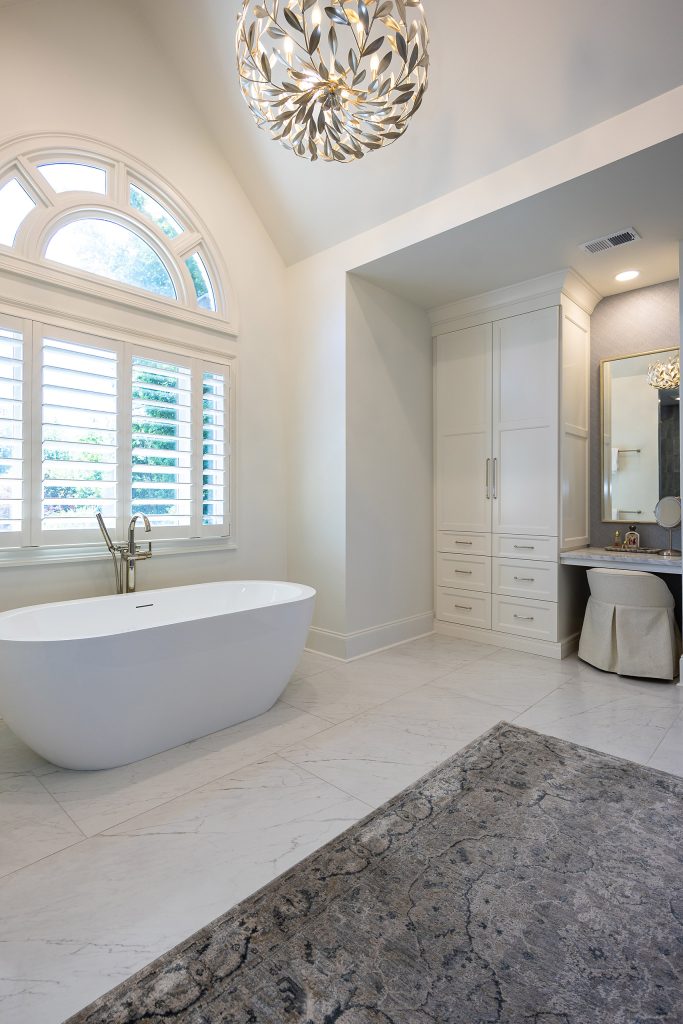 Master bath with a freestanding tub, arched window, glass shower in grey-blue tile, and a linen cabinet.