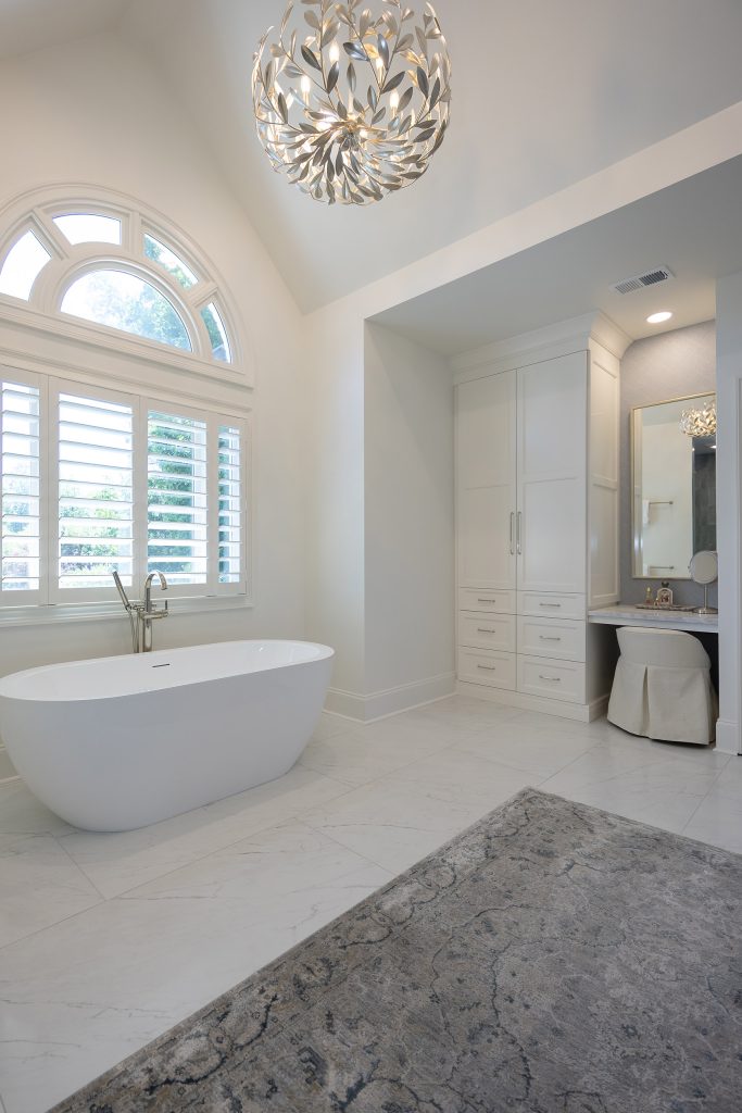 Master bath with a soaking tub, arched window, tall white linen cabinet, and a marble-topped makeup nook.
