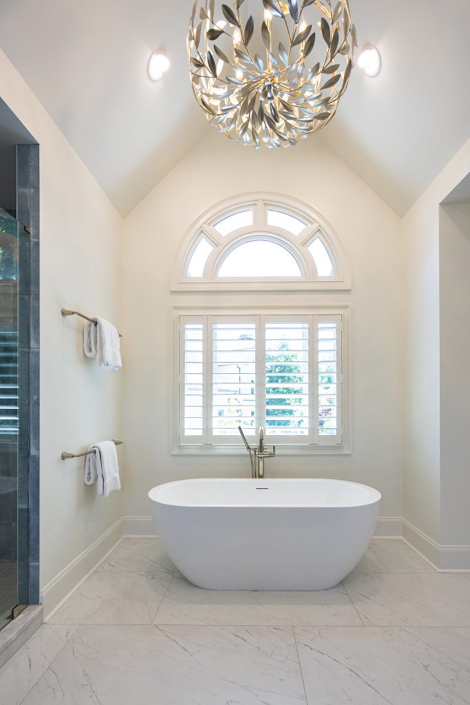 Freestanding tub below an arched Palladian window with a silver branch-and-leaf chandelier above.