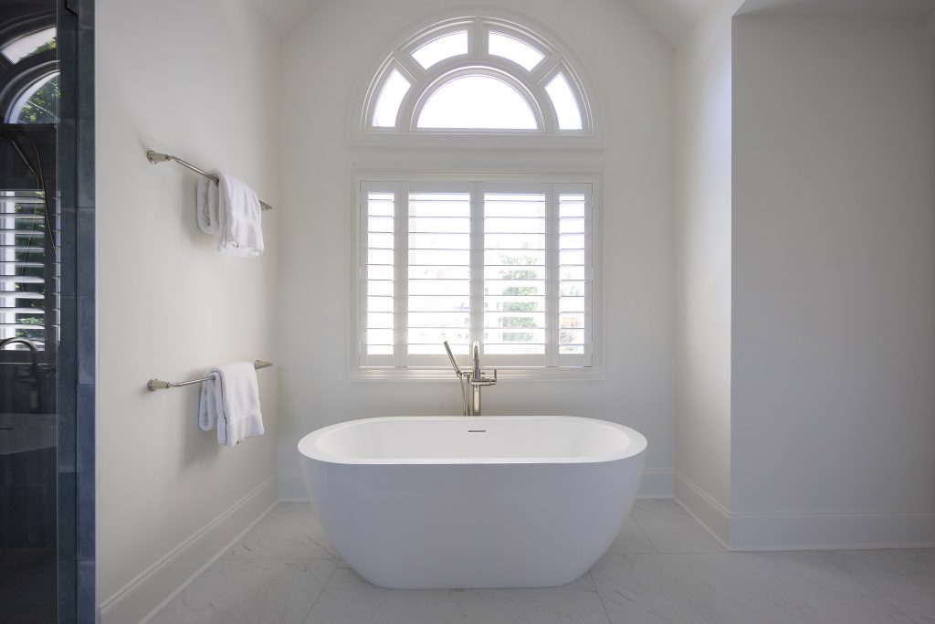 Freestanding tub beneath an arched Palladian window with plantation shutters and marble-look flooring.