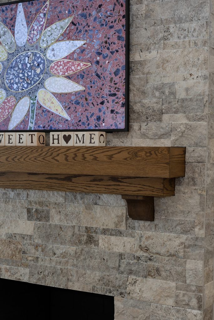 Detail of a stacked-stone fireplace with a chunky stained-oak mantel and a terrazzo sunflower panel above.